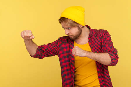 Determined hipster bearded guy in beanie hat, checkered shirt holding clenched fists up ready to boxing, punching with confident expression, fighting spirit. studio shot isolated on yellow, side viewの写真素材