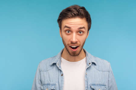 Portrait of astonished man in worker denim shirt looking at camera with open mouth, big eyes, feeling amazed shocked and happy about unexpected success. indoor studio shot isolated on blue backgroundの写真素材