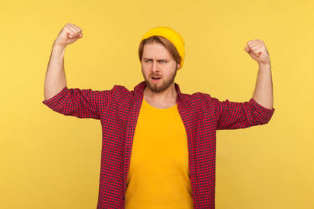 I am strong! Proud hipster bearded guy in beanie hat and checkered shirt showing biceps, feeling...