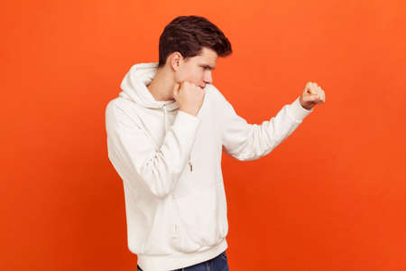 Male fighter in hoodie holding clenched fists up, ready to attack. Young man defending from bullies. Martial art school lessons. Indoor studio shot isolated on orange backgroundの写真素材