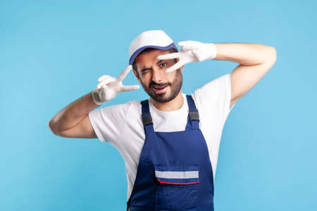 Positive handyman in overalls and hygiene gloves showing victory sign and winking playfully to camera, profession of service industry, courier delivery, housekeeping maintenance. studio shot isolatedの写真素材