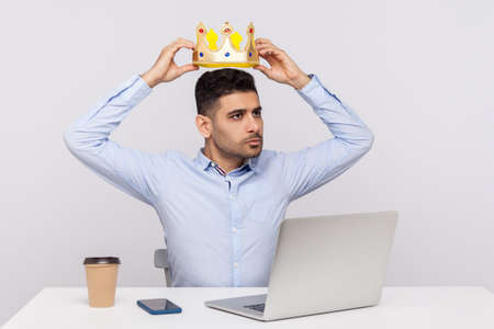 Proud authoritative businessman sitting in office workplace, wearing crown on head and looking away self-confident arrogant expression. indoor studio shot isolated on white backgroundの写真素材