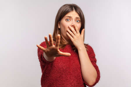 Portrait of panicked scared girl in shaggy sweater gesturing stop and looking surprised with frightened eyes, covering mouth not to shout from fear. indoor studio shot isolatedの写真素材