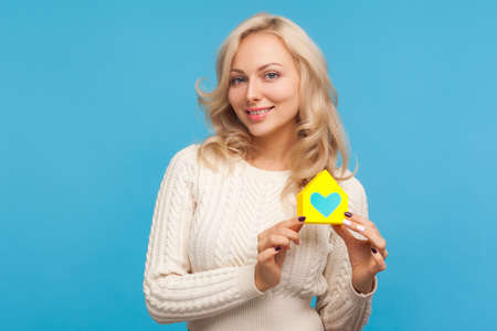 Happy smiling woman with curly blond hair holding paper house with painted heart and looking at camera, charity, shelter. Indoor studio shot isolated on blue backgroundの写真素材