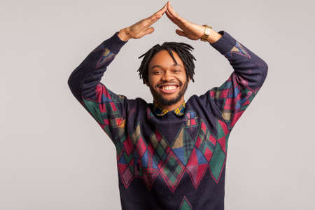 Extremely satisfied african man with dreadlocks showing house gesture holding hands above head with toothy smile on face, real estate agency. Indoor studio shot isolated on gray backgroundの写真素材