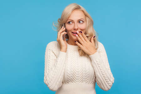 Surprised shocked blond woman in knitted sweater talking phone, discussing friends, amazed with heard news, gossip. Indoor studio shot isolated on blue backgroundの写真素材