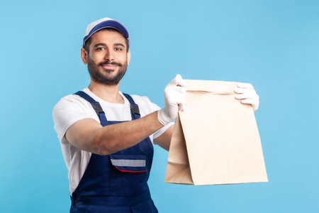 Take it! Portrait of friendly smiling handyman in overalls and gloves giving ordered parcel. Courier delivering food in paper bag, post mail services. indoor studio shot isolated on blue backgroundの写真素材