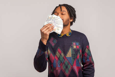 Happy satisfied african guy with dreadlocks and beard sniffing big batch of money enjoying big...