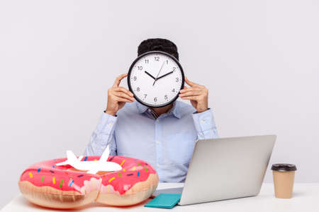 Time to rest! Unknown man hiding face behind clock, sitting in office workplace with rubber ring...
