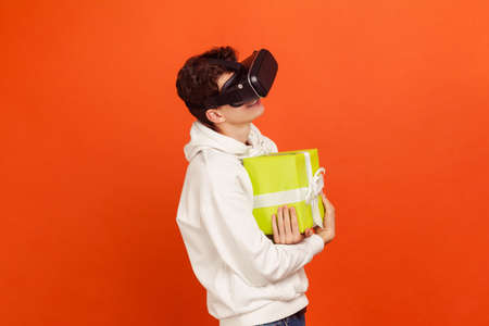 Cheerful satisfied teenager in casual style sweatshirt and virtual reality glasses hugging present...