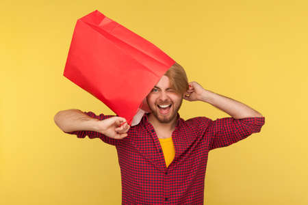 Crazy positive shopper guy in checkered shirt wearing shopping bag on his head, having fun with...