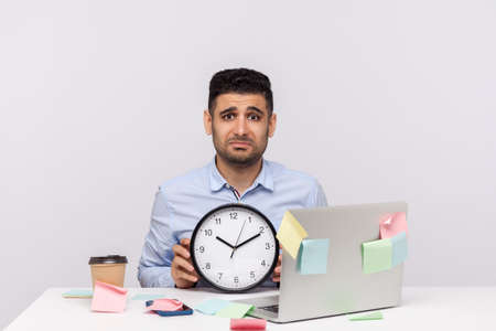 Overtime work. Unhappy exhausted man employee sitting in office workplace, holding clock and frowning upset, sticky notes all around reminding of deadline. studio shot isolated on white backgroundの写真素材