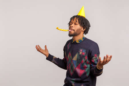 Pleased satisfied african man with dreadlocks in party hat having fun and enjoying blowing party horn with closed eyes, birthday celebration. Indoor studio shot isolated on gray backgroundの写真素材