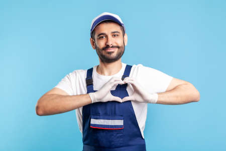 Love our customers! Handyman in overalls and hygiene gloves showing heart gesture and smiling...
