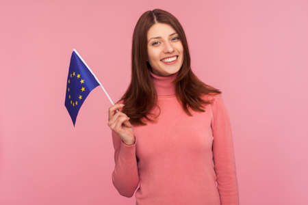 Happy patriotic woman with brown hair holding eu flag in hand looking at camera with toothy smile, patriotism, flag day celebration. Indoor studio shot isolated on pink backgroundの写真素材