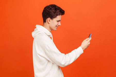Profile of happy smiling teenager in casual hoodie holding mobile phone in hands chatting via video...