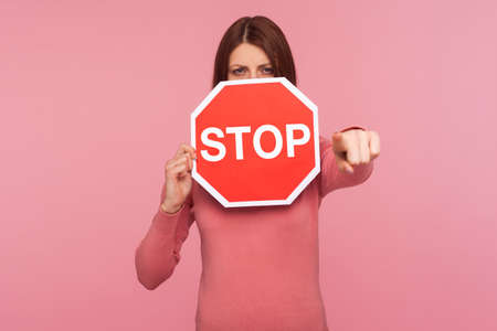 Angry strict woman in pink sweater holding stop warning sign and pointing finger at you, access...