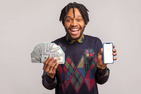 Extremely happy african man with dreadlocks satisfied holding mobile phone and fan of dollars in hands with toothy smile, online earning. Indoor studio shot isolated on gray backgroundの写真素材