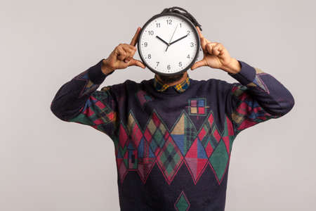 Time to work! African man hiding face behind big wall clock, afraid of deadline, wasting time, being late. Indoor studio shot isolated on gray backgroundの写真素材