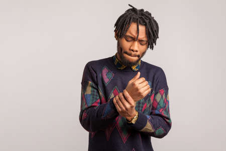 Frustrated sad african man with dreadlocks grimacing touching his wrist, feeling sudden acute pain,...