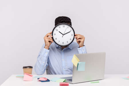 Overtime work. Anonymous man employee sitting in office workplace, hiding face behind clock, sticky notes all around reminding of deadline, stressful job. studio shot isolated on white backgroundの写真素材