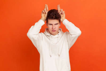 Arrogant angry teenager in fashionable white hoodie showing horns, holding fingers near head,...