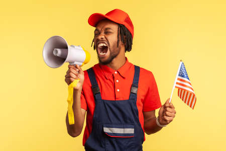 Angry nervous afro-american worker in uniform with beard and dreadlocks holding usa flag screaming at loudspeaker, protesting. Indoor studio shot isolated on yellow backgroundの写真素材