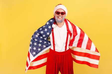 Happy patriotic grey bearded man in santa claus hat and sunglasses standing wrapped in usa flag and looking at camera with toothy smile. Indoor studio shot isolated on yellow backgroundの写真素材