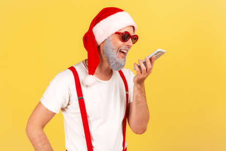 Smiling happy grey bearded man in santa claus hat and stylish glasses holding smartphone near mouth talking to voice assistant, leaving voice message. Indoor studio shot isolated on yellow backgroundの写真素材