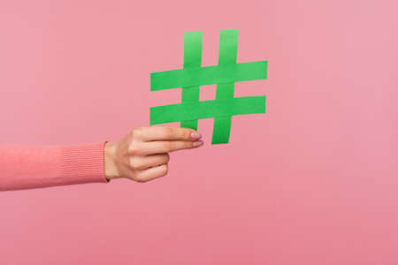 Join popular blog! Closeup of female hand holding green hashtag sign, sharing tagged message, recommending follow trends. Indoor studio shot isolated on pink backgroundの写真素材