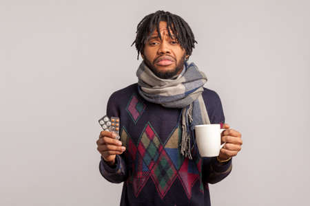 Deeply frustrated african guy with dreadlocks in checkered scarf holding cup of tea and pills in hands sadly looking at camera, cold and flu. Indoor studio shot isolated on gray backgroundの写真素材