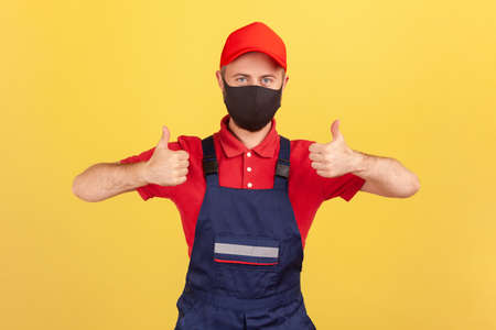 Positive crafts man in red and blue uniform and protective facial mask showing thumbs-up, satisfied...