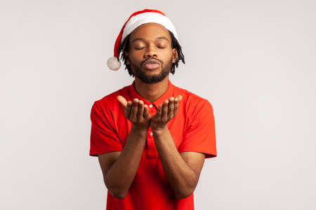 Positive handsome afro-american man with beard and dreadlocks in santa claus hat sending air kisses to camera closing eyes. Indoor studio shot isolated on gray backgroundの写真素材