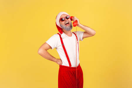 Friendly optimistic man in sunglasses, pants with suspenders and santa claus hat talking on landline telephone holding handset, greeting with holidays. Indoor studio shot isolated on yellow backgroundの写真素材