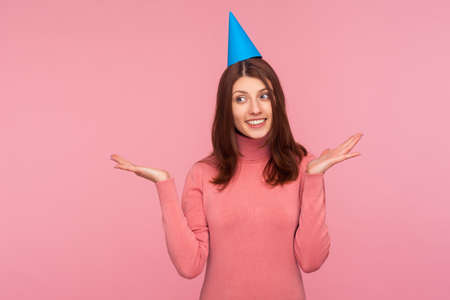 Joyful pretty brunette woman in funny party cap smiling and spreading hands saying surprise, festive...