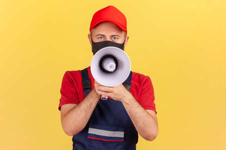 Craftsman in uniform and facial protective mask speaking holding loudspeaker near mouth, talking...
