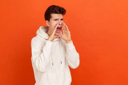 Profile portrait of hysterical young man loudly yelling in panic, holding hands near his mouth, teenager protest. Indoor studio shot isolated on orange backgroundの写真素材