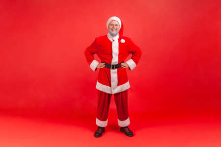 Full length portrait of positive happy aged man in santa claus costume standing holding hands akimbo, preparing for winter holidays and toothy smile. Indoor studio shot isolated on red backgroundの写真素材