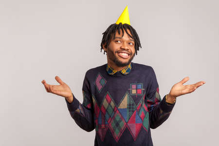 Happy surprised african man with beard and dreadlocks in yellow cone hat on head spreading his hands, wondered with party, personal entertainment. Indoor studio shot isolated on gray backgroundの写真素材