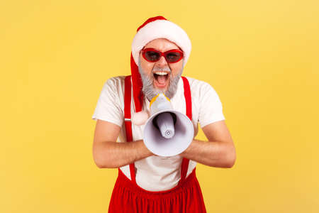 Excited emotional gray bearded man in santa claus costume screaming at loudspeaker, congratulates with holidays, fancy party. Indoor studio shot isolated on yellow backgroundの写真素材
