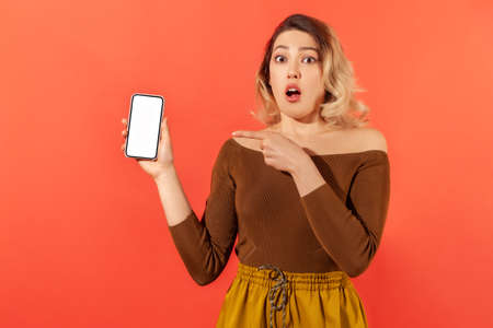 Shocked surprised woman pointing finger at smartphone with white empty screen, looking with amazed...