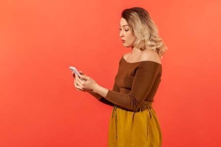 Profile portrait attractive woman looking at smartphone display with amazed expression, reading comments in social networks, sudden news. Indoor studio shot isolated on orange backgroundの写真素材