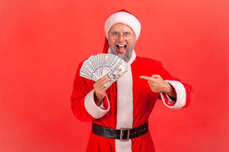 Extremely happy excited man in santa claus costume pointing finger at dollar banknotes he holding in...