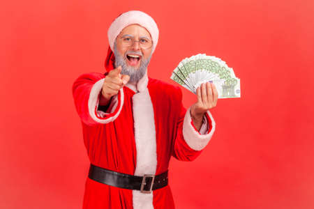 Extremely happy excited santa claus holding fun of euros and pointing finger at you looking at...