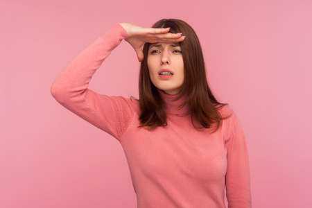 Curious brunette woman in pink sweater looking far away with hand over head, trying to see...