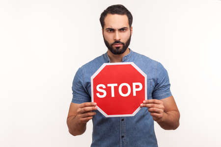 Concerned nervous bearded man in blue shirt holding red stop sign looking at camera with serious expression. Indoor studio shot isolated on white backgroundの写真素材