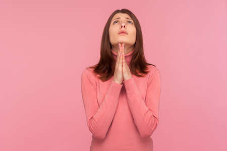 Lonely upset brunette woman looking up holding hands together, praying God, asking for health and support pleading for healing and forgiveness. Indoor studio shot isolated on pink backgroundの写真素材