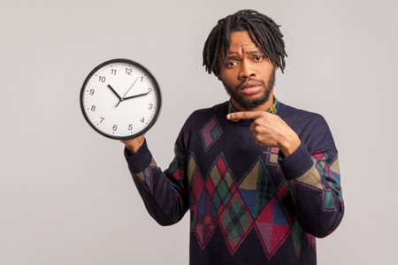 Nervous african man with beard and dreadlocks pointing finger on clock in his hand with serious face, time management, schedule. Indoor studio shot isolated on gray backgroundの写真素材