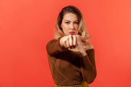 Boxer! Portrait of young woman with blonde hair in brown blouse punching fist to fight, staring...