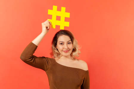 Attractive positive woman blogger holding yellow hashtag sign on her head and smiling at camera, tagging posts on internet. Indoor studio shot isolated on orange backgroundの写真素材
